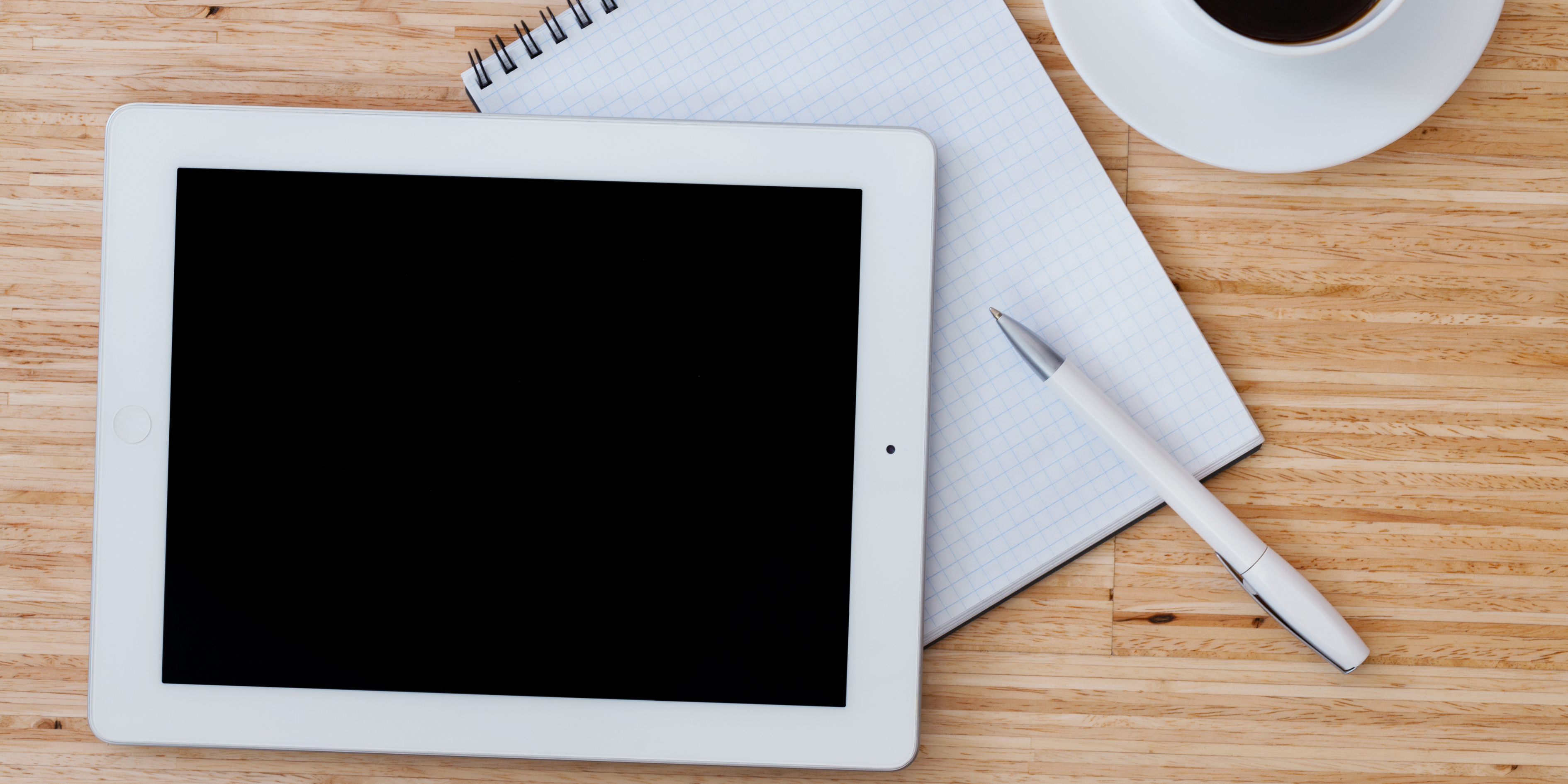 White tablet with a black screen placed on a wooden desk beside a notepad, pen, and cup of coffee — representing a device that may not be charging.